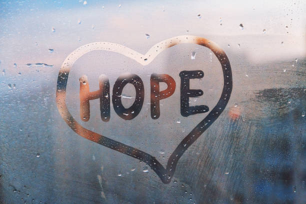 Handwritten word Hope in shape heart on misted glass on window flooded with raindrops on blue cloudy background